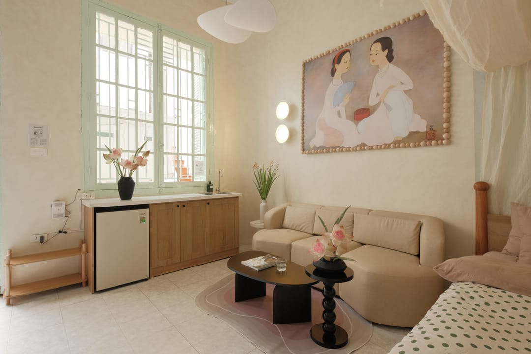 The sitting corner of Studio 1A: a beige sofa beneath the áo dài painting, a compact wooden kitchenette and mini-fridge below a tall mint-green shuttered window