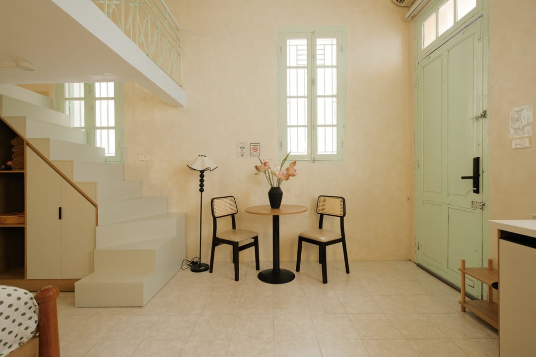 A small round table with two black cane chairs beside a white staircase climbing to the mezzanine, next to the tall mint-green entrance door of Studio 1A