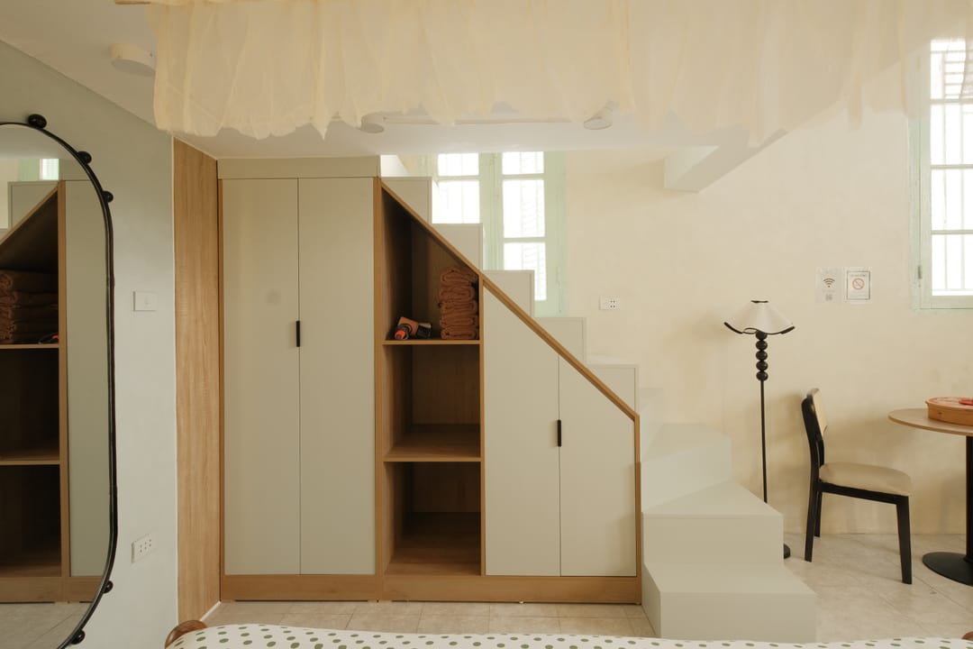 An oak wardrobe and open shelves built into the space under the white staircase in Studio 1A, with an arched mirror on the left and a floor lamp beside a small table