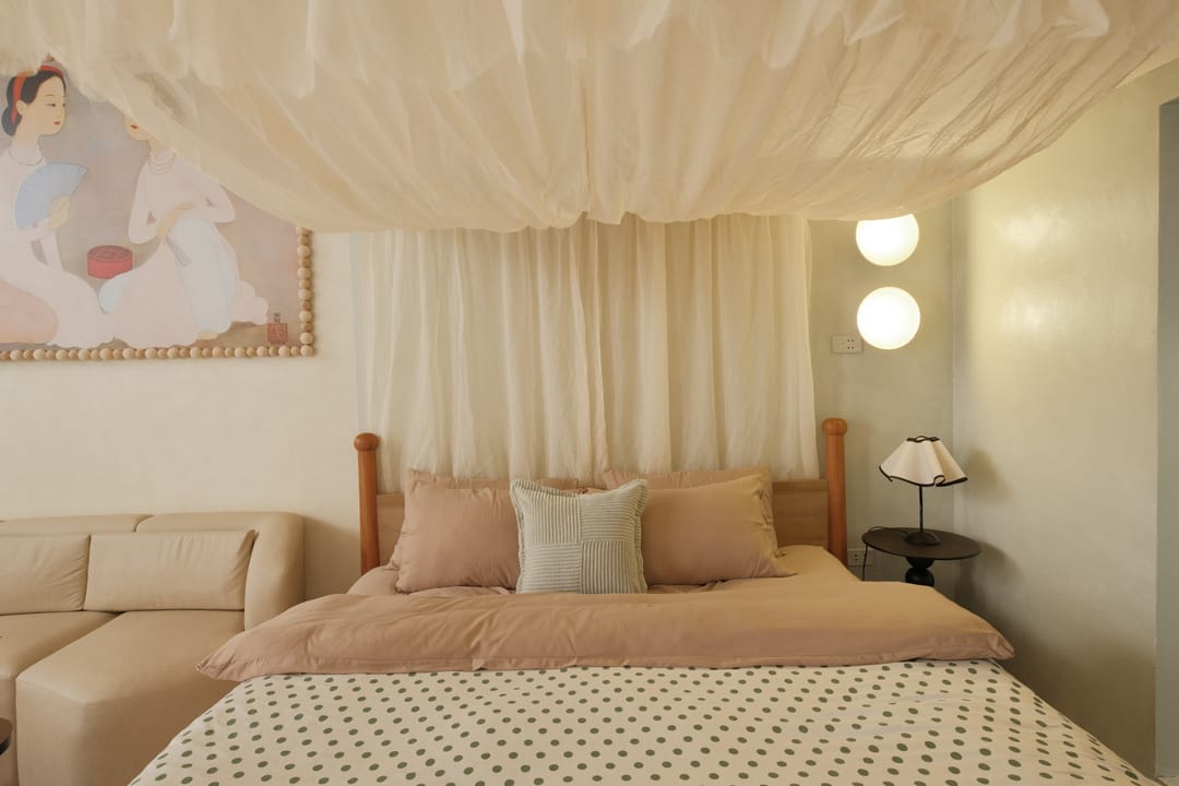 Close view of the canopied king bed in Studio 1A with blush pillows and a polka-dot cover, the áo dài painting on the left and two globe wall lamps on the right