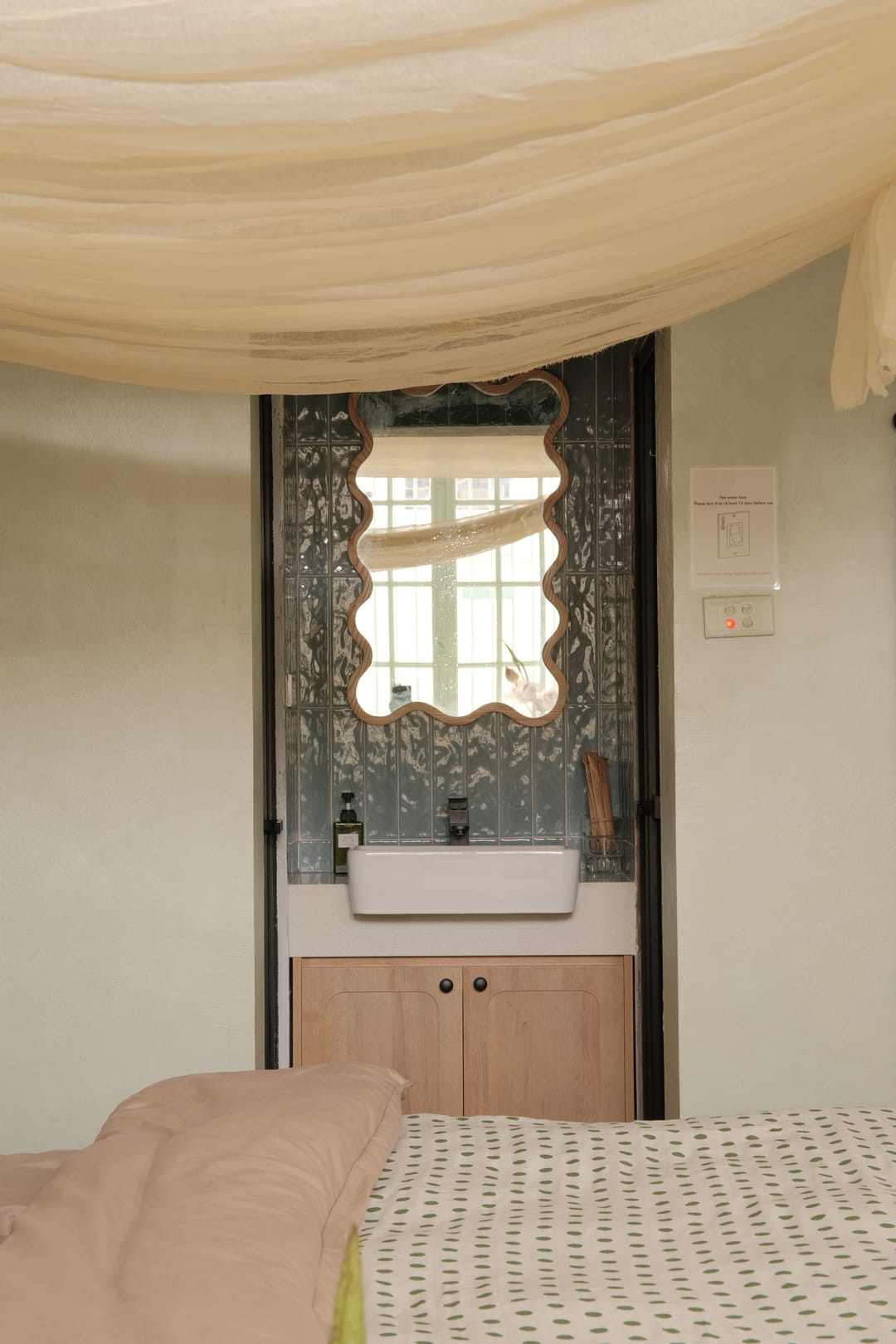 A washbasin set into a niche beside the bed in Studio 1A, a wavy wooden-framed mirror above it framed by black-edged glazed doors, with the muslin canopy overhead