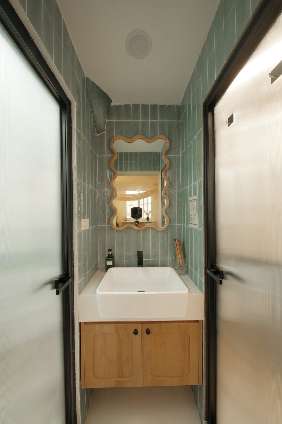 The bathroom of Studio 1A: sage-green tiles, a wavy wooden-framed mirror over a white basin on an oak vanity, between two black-framed frosted-glass doors