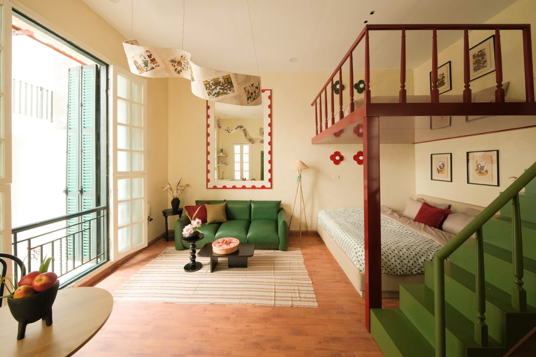The sitting end of Studio 2A: green-shuttered French doors, a mirror in a red scalloped frame above a green leather sofa, and a folk-art printed pendant shade over the coffee table
