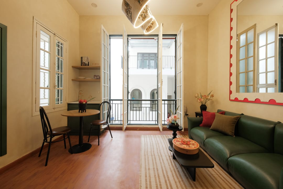 Open white French doors onto a small balcony over a courtyard in Studio 2A, with a round dining table and bentwood chairs, a green sofa and the red scalloped mirror