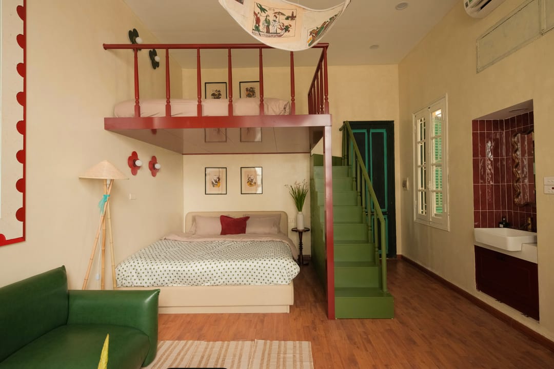 Studio 2A from the sofa: a green staircase to the red-railed mezzanine bed, framed folk prints, a green door and a tiled bathroom nook with a basin on the right