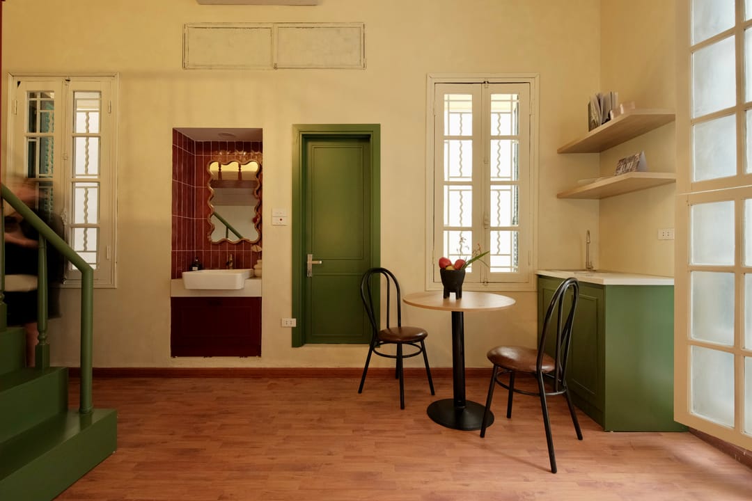 The dining corner of Studio 2A: a round wooden table with two bentwood chairs, a green door, a green kitchenette and a maroon-tiled bathroom niche with a wavy mirror and basin