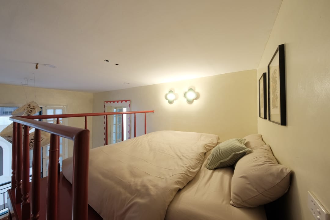 The mezzanine bed of Studio 2A behind red railings, with two flower-shaped wall lamps, framed folk prints and a view down toward the balcony window