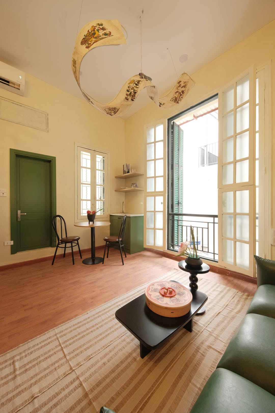 A tall view of Studio 2A's living room: a folk-art printed pendant shade curling overhead, a green door, the dining nook, French balcony doors and a green leather sofa