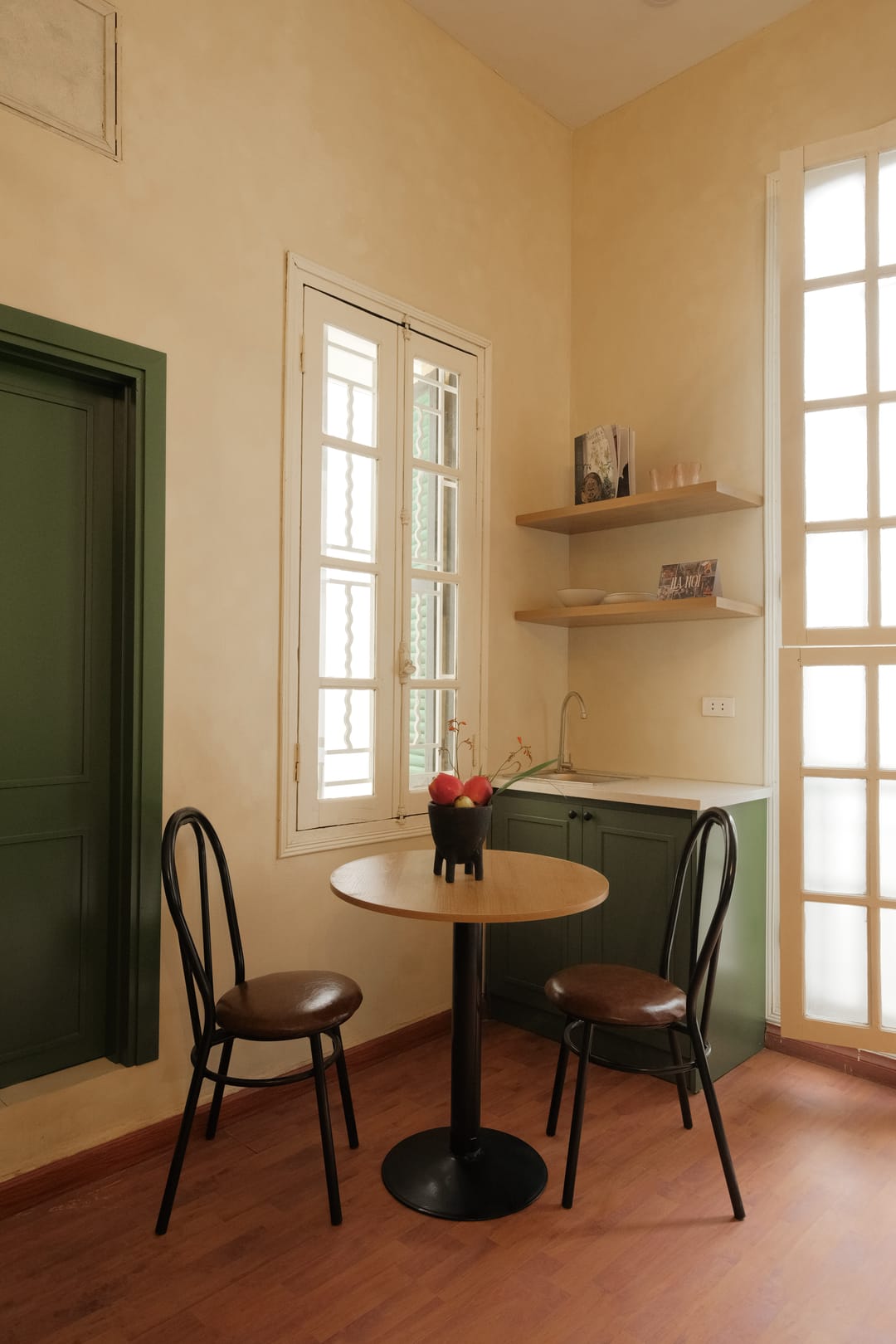 The dining nook of Studio 2A: a round wooden table with two black bentwood cane chairs, a green door and a green kitchenette with open shelves below a shuttered window