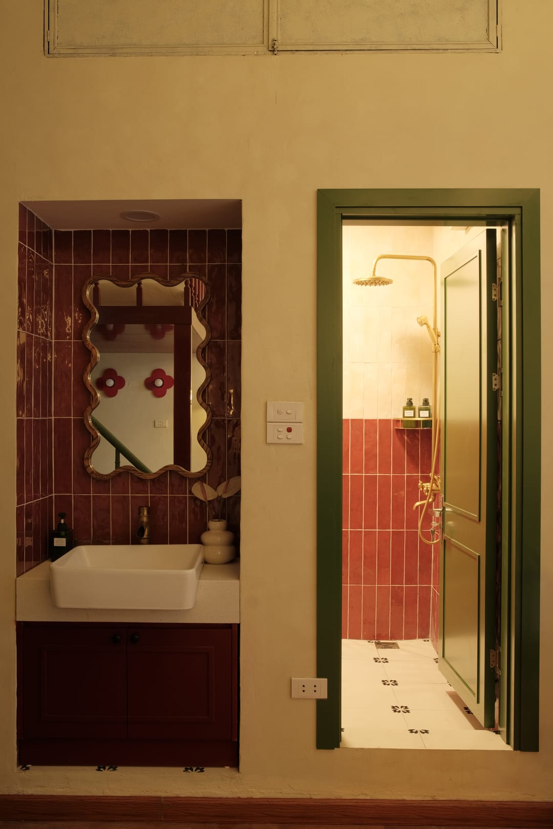 The bathroom of Studio 2A: a maroon-tiled niche with a wavy gold-framed mirror, red flower hooks and a white basin, beside an open green door into the tiled shower