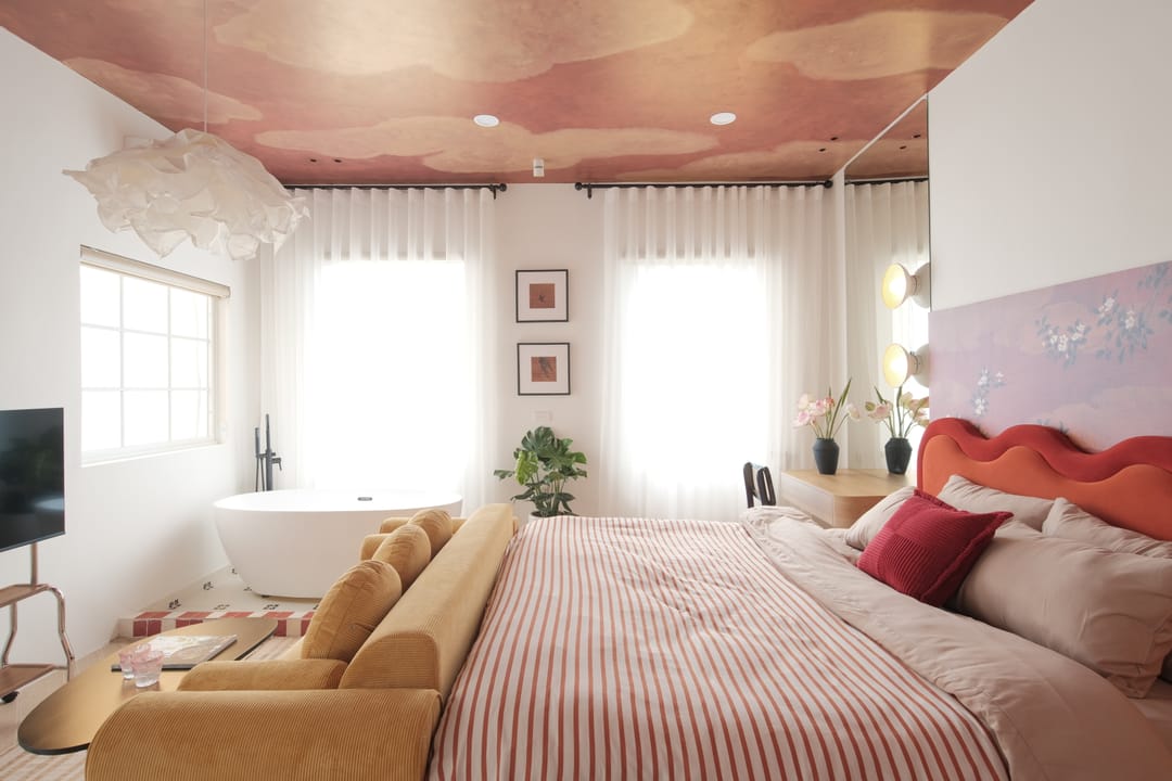 Studio 3A: a freestanding bathtub in the light of curtained windows, a television, and the red bed with striped bedding beneath the cloud ceiling