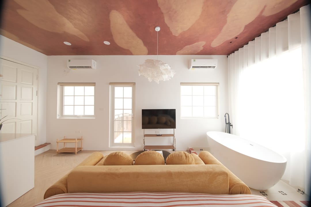 A wide view of Studio 3A over the mustard sofa: tall windows, a wall-mounted television, the freestanding bath on the right and the red bed, under the painted cloud ceiling