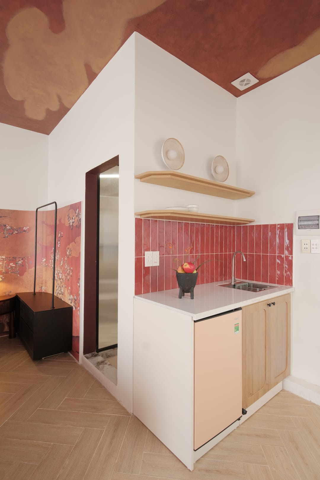 The kitchenette in Studio 3A: a pink-tiled splashback, open wooden shelves, a small fridge and a bowl of fruit, beside a glazed door to the shower under the cloud ceiling