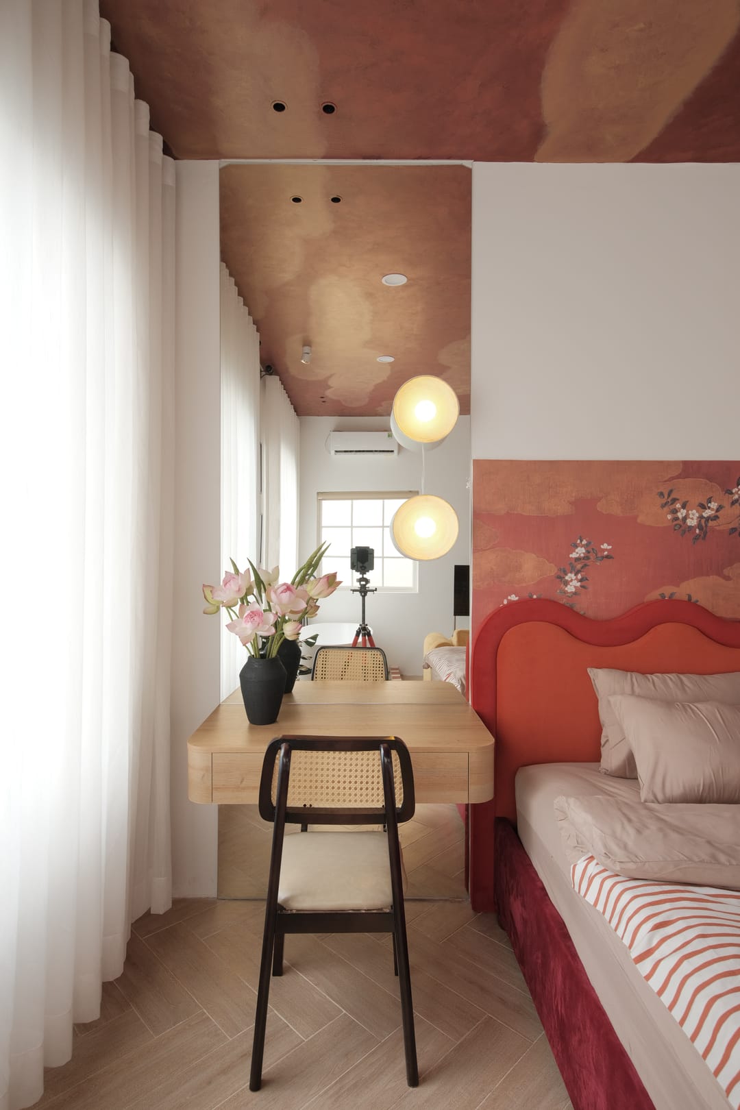 A wooden desk with a cane chair and a vase of pink lotus in Studio 3A, lit by two glass globe pendants beside sheer curtains, with the blossom wall and red bed on the right