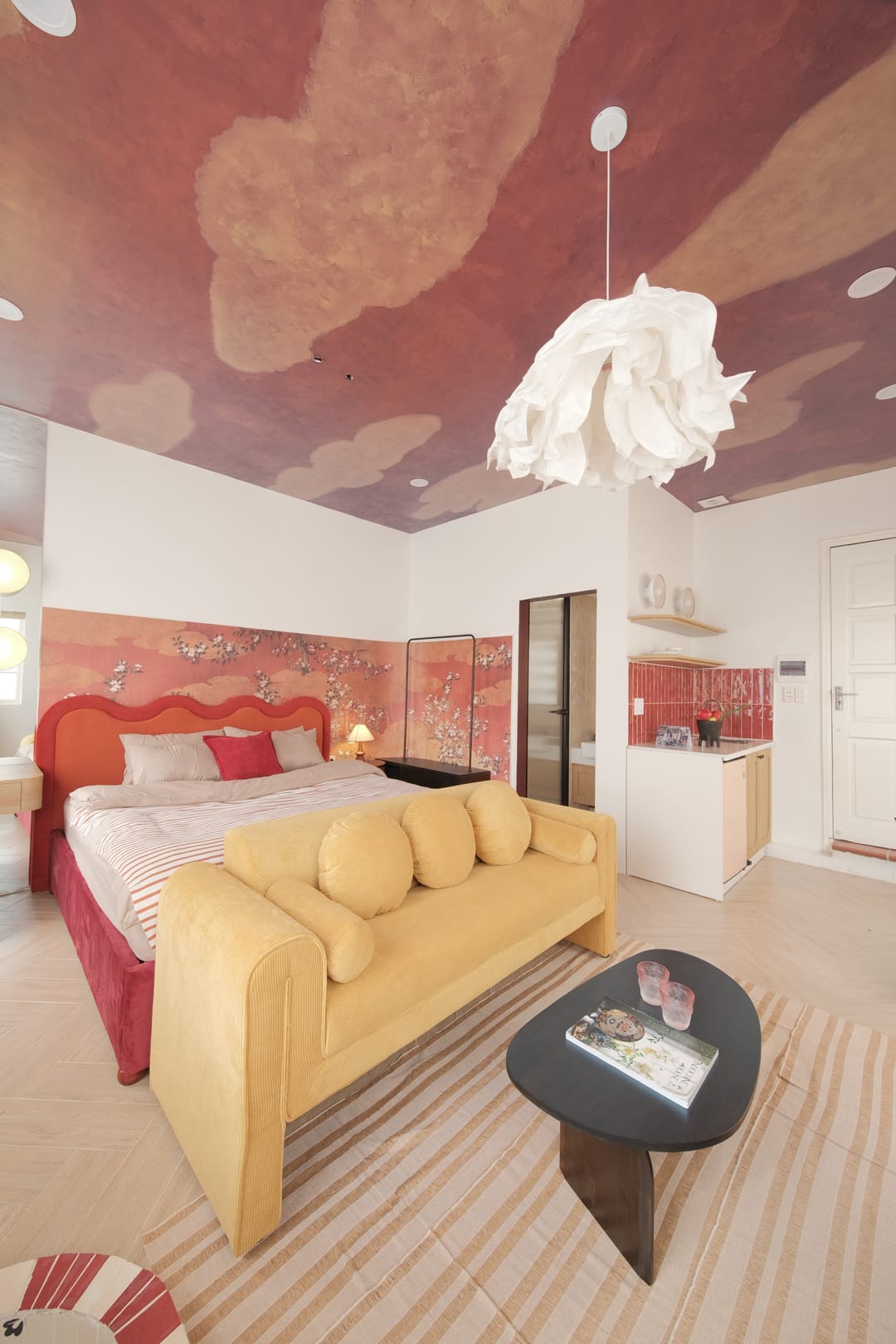 Studio 3A from the doorway: the red bed and mustard sofa on a striped rug, the pink blossom wall and kitchenette, beneath a sculptural white pendant and the cloud ceiling