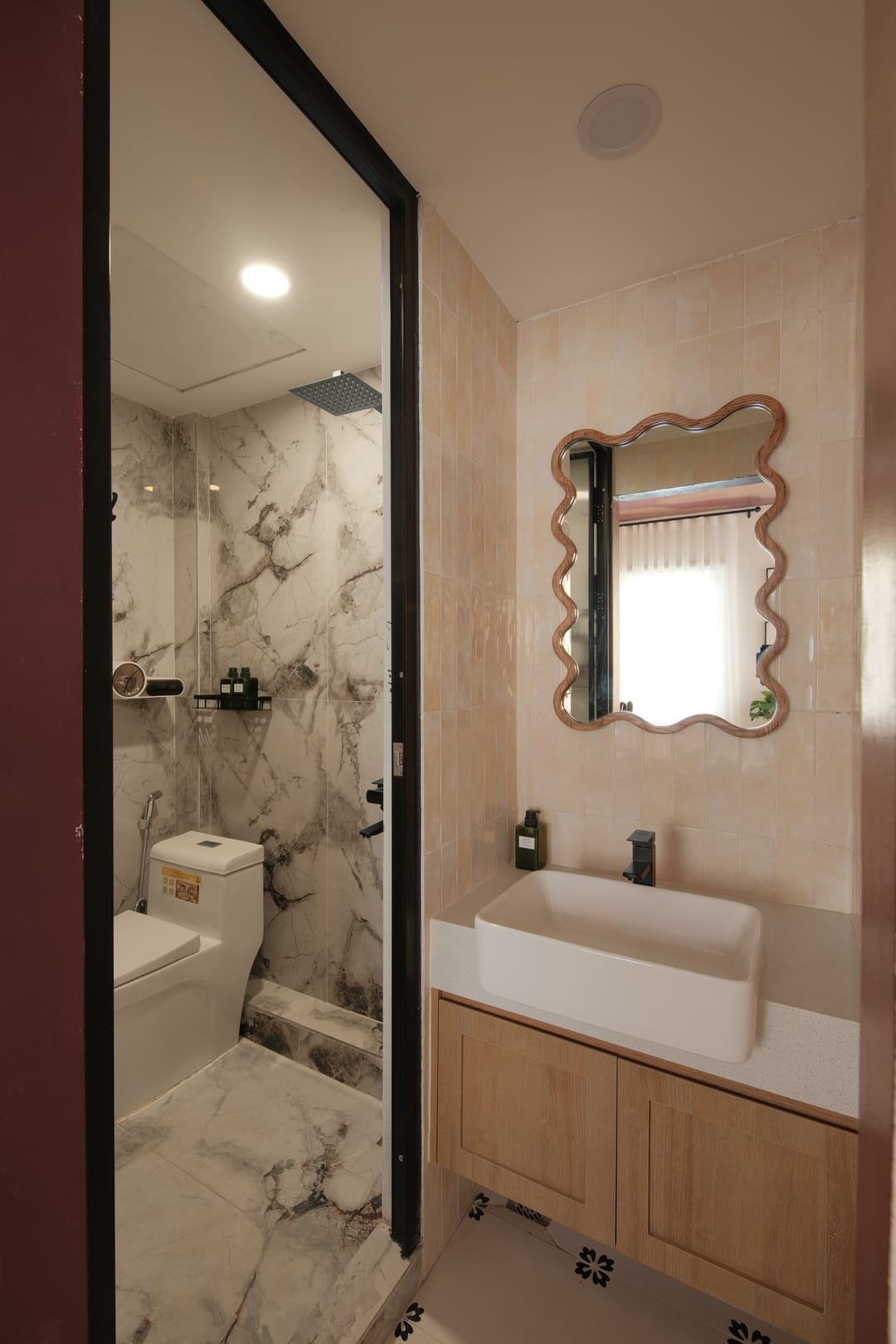 The bathroom of Studio 3A: a marble-lined walk-in shower behind a black-framed glass door, a wavy wooden-framed mirror over a white basin and an oak vanity