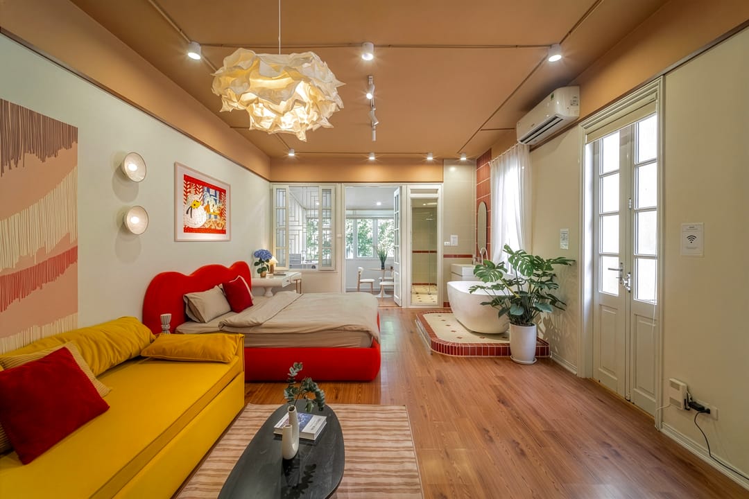 Studio 4A under a cloud-like pendant: the red bed, yellow sofa bed, a freestanding bath on a tiled plinth by the French doors, and a monstera plant