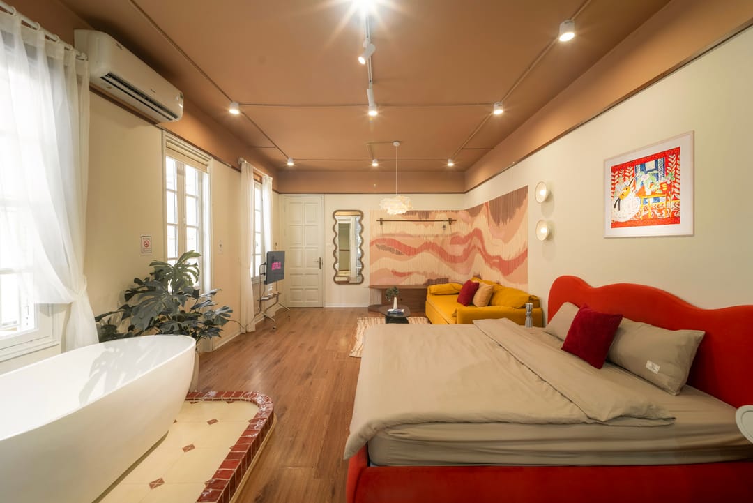 A wide view of Studio 4A: the freestanding bath on a tiled plinth by the window, a television, a wavy mirror and white door, the coral wave wall, yellow sofa and red bed with the folk-art print