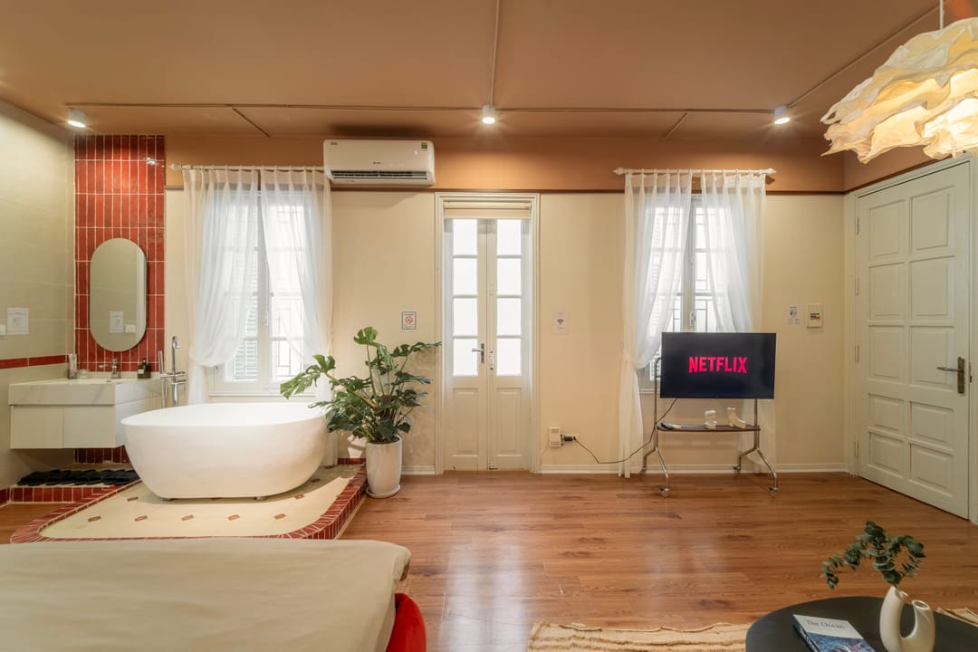 The bathing end of Studio 4A: a red-tiled washstand with an oval mirror, a freestanding bath between two curtained windows, French doors and a television showing Netflix on a wheeled stand