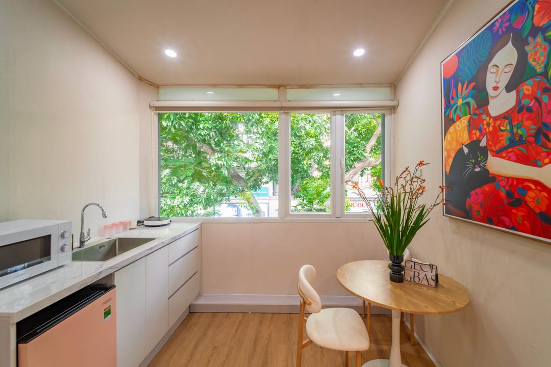 The kitchen and dining corner of Studio 4A: a white kitchenette with a microwave and pink mini-fridge, a window full of leaves, and a round table below a painting of a woman in red holding a black cat