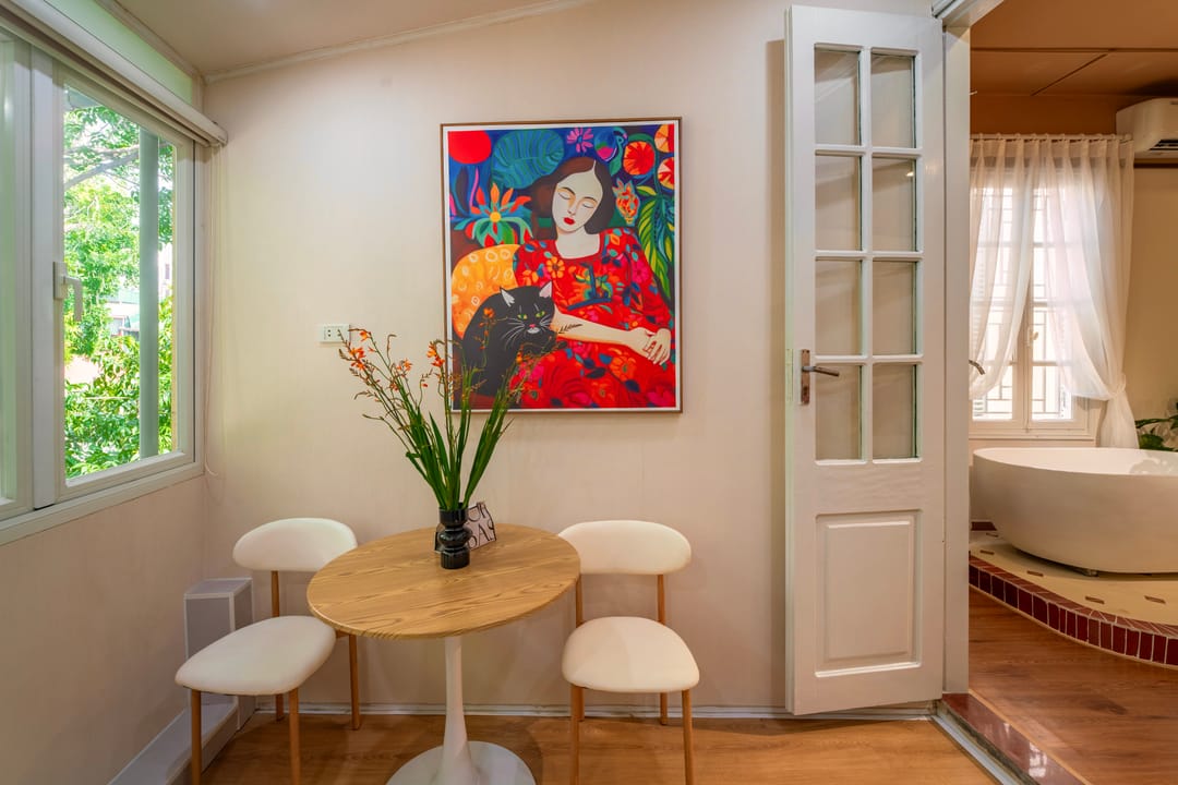 The dining nook of Studio 4A: a round table with cream stools below a bright painting of a woman holding a black cat, beside a leafy window and a white door open to the bathtub