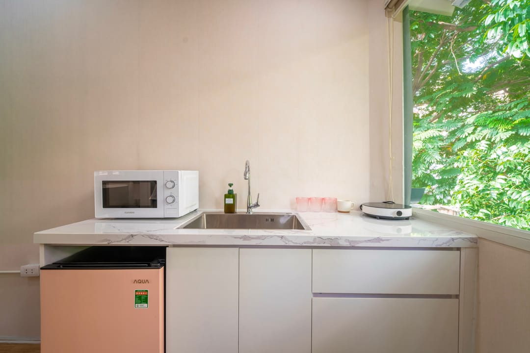 Close view of the kitchenette in Studio 4A: a white marble counter with a microwave and sink, a pink mini-fridge below and a window filled with green leaves
