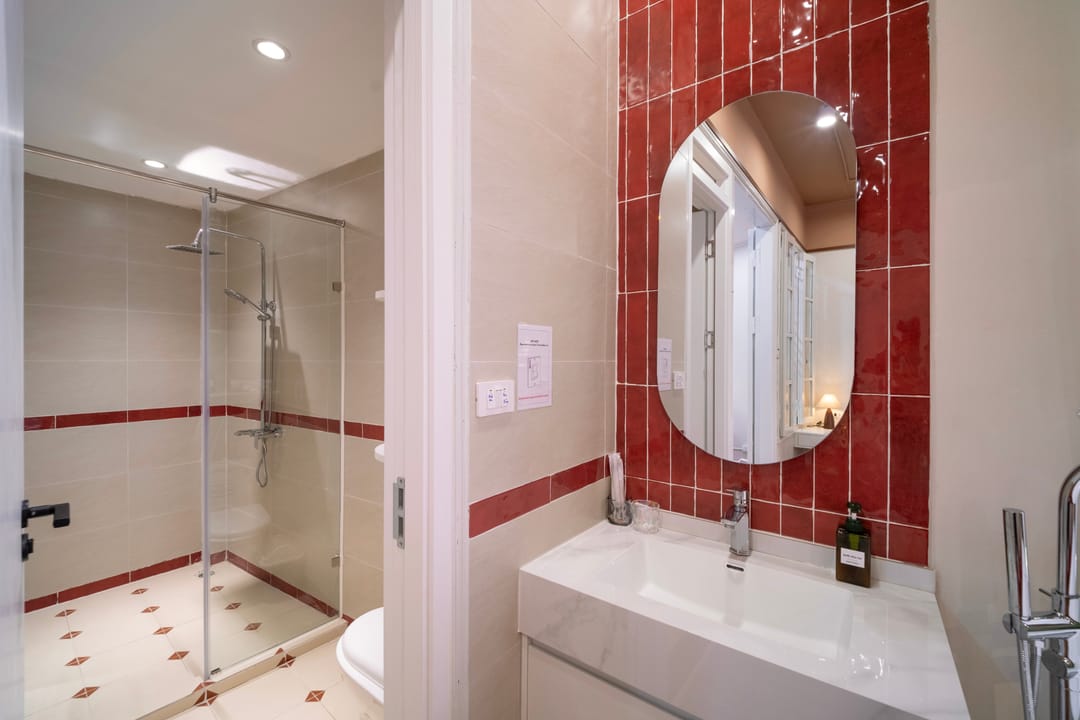 The bathroom of Studio 4A: a glass shower cubicle on the left, a red-tiled wall with an oval mirror over a white basin, and a floor-standing tap on the right
