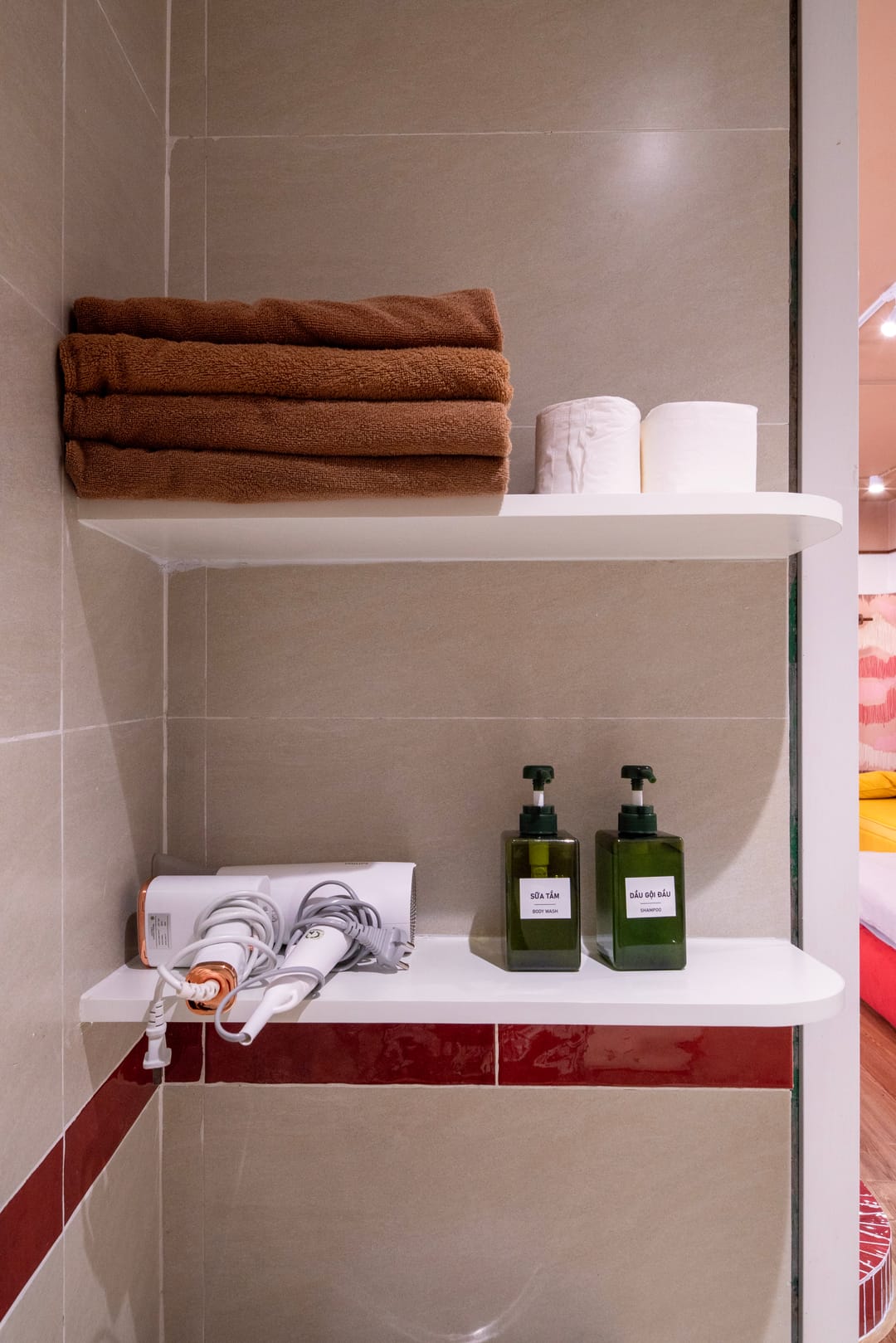 A shelf in Studio 4A's bathroom: folded brown towels and toilet rolls above a hairdryer and two green pump bottles of body wash and shampoo, against beige tile