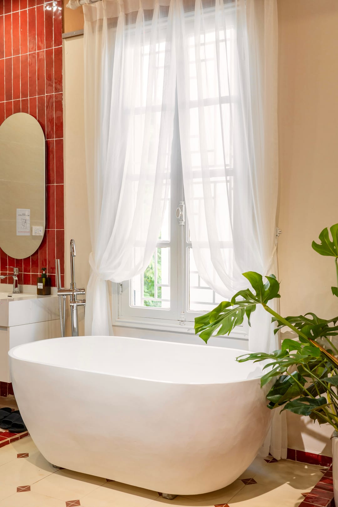 A freestanding white bath by a tall window with sheer curtains in Studio 4A, beside a red-tiled wall with an oval mirror and a large monstera plant