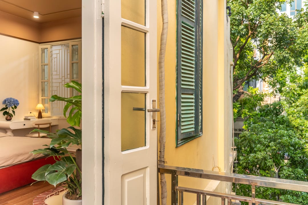 The balcony of Studio 4A: a white French door open from the room onto a small railed balcony, with green window shutters, tree canopy and the street below